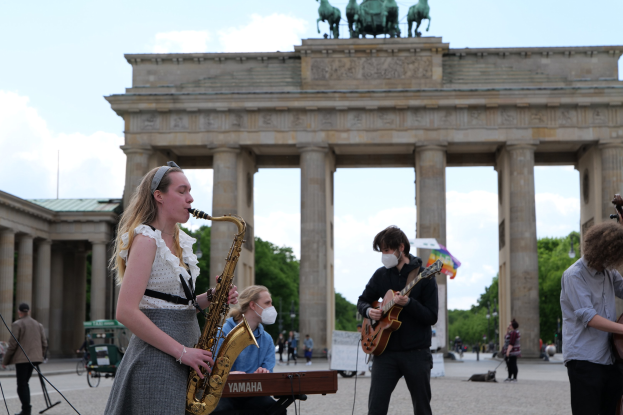 Eine Gruppe von Menschen, die Musikinstrumente vor dem Brandenburger Tor in Berlin spielen, darunter eine Frau an der Saxophon, ein Mann an der Gitarre und eine maskierte Person, mit Bäumen, Gebäuden, Säulen, Statuen und bewölktem Himmel im Hintergrund.