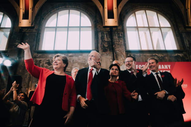 Eine Gruppe von Labour Party-Mitgliedern, die feierlich klatschen und vor einer Menge mit einem Banner und beleuchteten Fenstern im Hintergrund stehen.