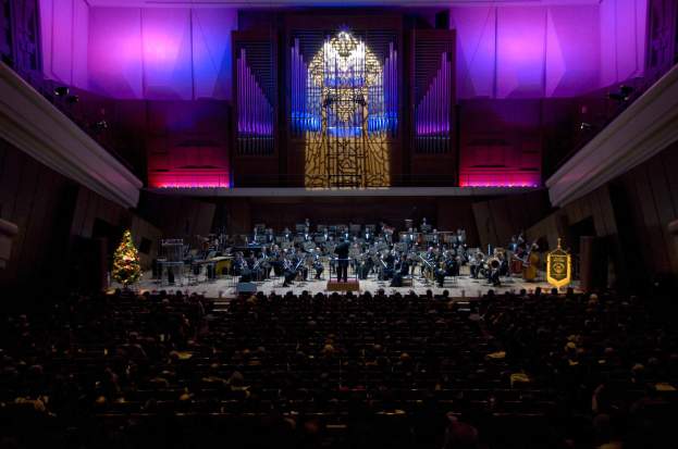Großer Saal mit Menschen bei einem Konzert, Musiker auf der Bühne mit Instrumenten, andere sitzend, Weihnachtsbaum im Hintergrund, Wände mit Lichtern und Schmuck dekoriert.