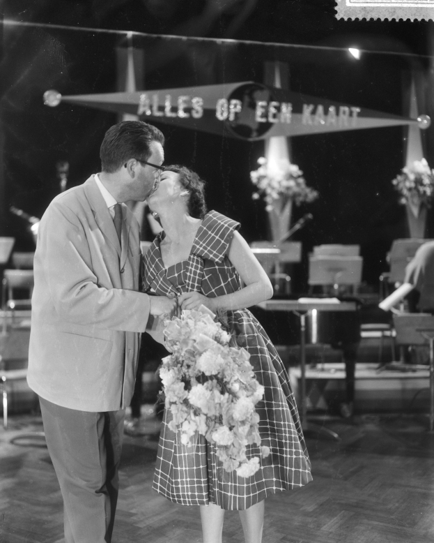 Ein Mann und eine Frau, die sich auf einer Bühne umarmen und küssen, wobei die Frau einen Blumenstrauß hält.