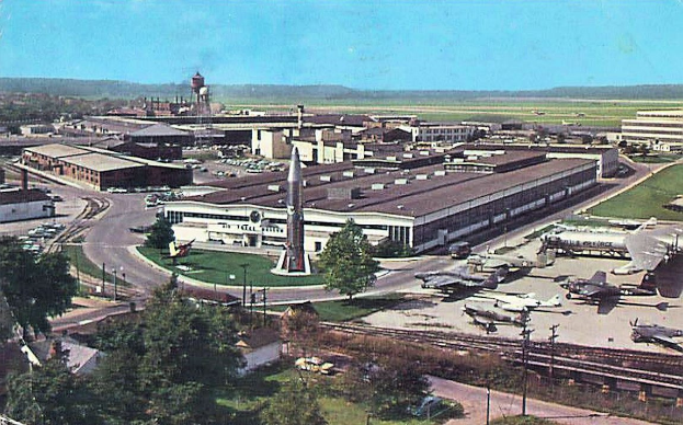 Alte Schwarz-Weiß-Airport-Szene mit geparkten Propellerflugzeugen, Terminalgebäuden, Bäumen, Straßen, Fahrzeugen, Versorgungsmasten, Gras, Bahnschienen, fernen Türmen, Hügeln und einem klaren Himmel, mit Untertext am unteren Rand.