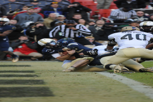 Ein Fussballspieler wird von einer Gruppe von Verteidigern auf einem Rasenfeld getackelt, mit Zuschauern und Kamerahaltern im Hintergrund, alle Spieler tragen Helme.