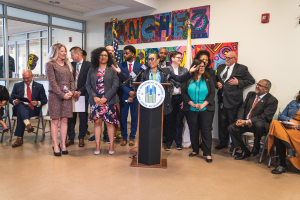 Gruppe von Menschen in einem Raum mit einem Podium, Mikrofon und einer Tafel mit dem Text "Sancho-Stadtrat tritt dem Stadtrat bei"; einige sitzen auf Stühlen und andere stehen, mit Flaggen und einem Feuerlöscher an der Rückwand.