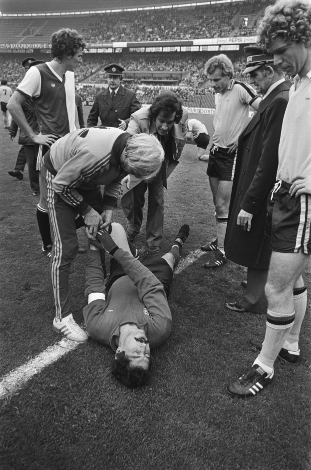 Schwarz-weißes Bild eines Fußballspielers, der auf dem Boden liegt und von einer Gruppe Männer umgeben ist, mit einer Menschenmenge im Hintergrund der Stadion.