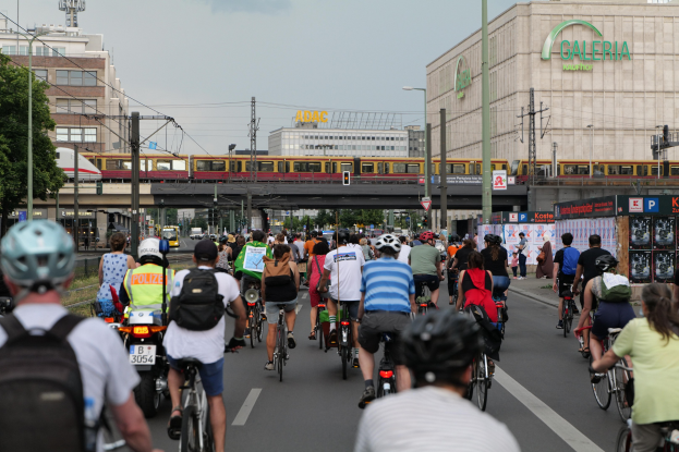 Eine Gruppe von Menschen, die Fahrräder auf einer Straße mit hohen Gebäuden fahren, wobei einige Helme und Taschen tragen, und ein Zug auf einem Bahngleis im Hintergrund.
