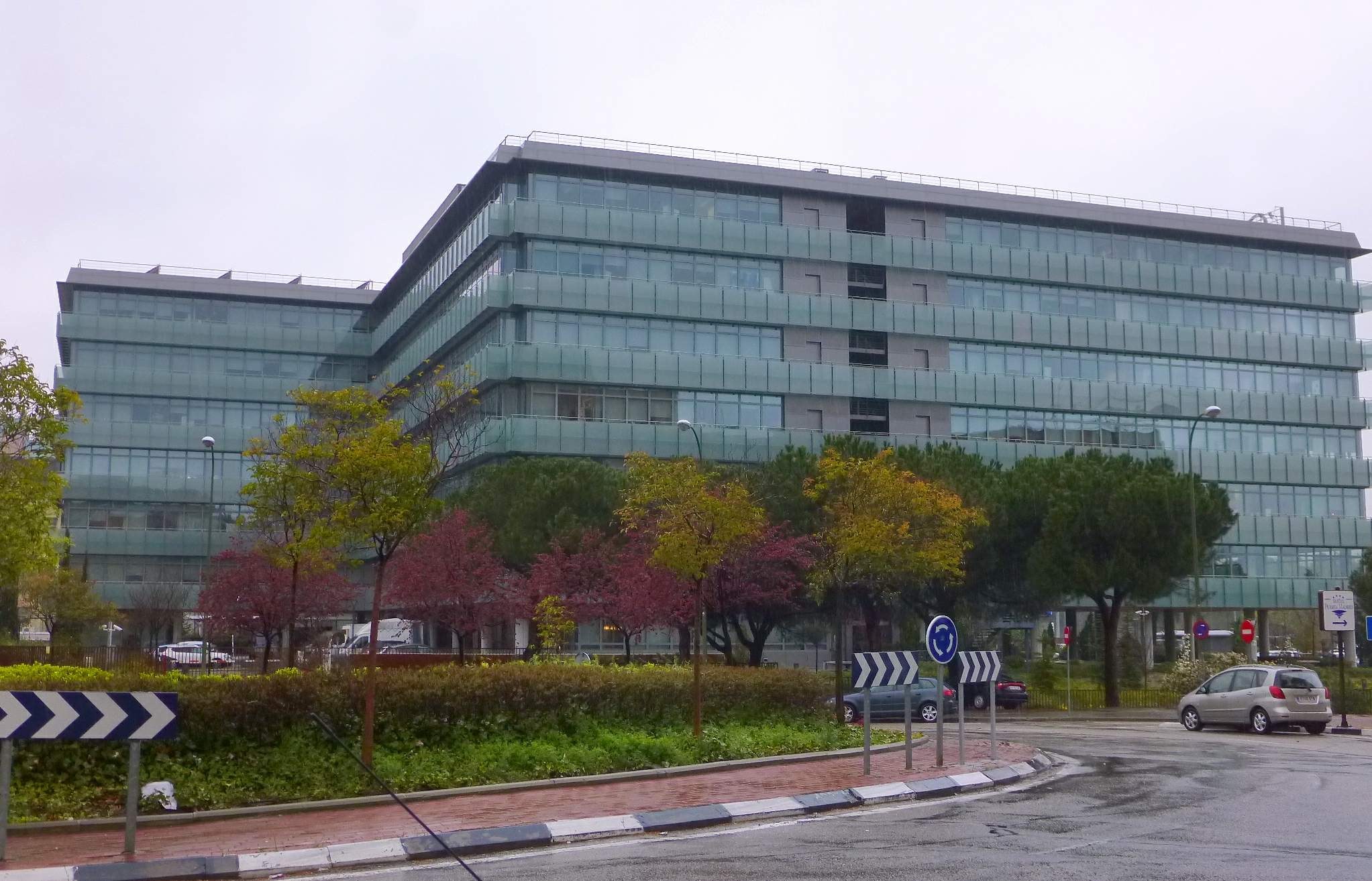 Großes modernes Gebäude mit zahlreichen Fenstern, identifiziert als Hauptquartier der Europäischen Zentralbank, umgeben von Grünflächen, Straßeninfrastruktur und Fahrzeugen unter einem bewölktem Himmel.