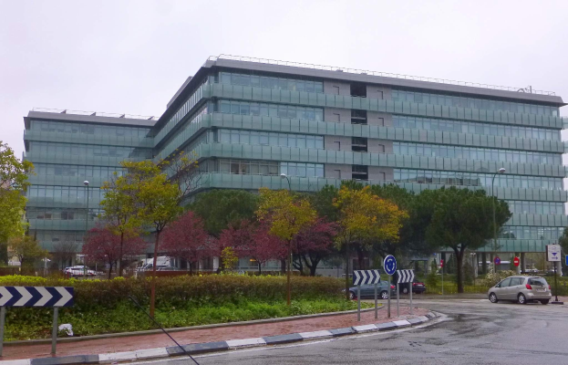 Großes modernes Gebäude mit zahlreichen Fenstern, identifiziert als Hauptquartier der Europäischen Zentralbank, umgeben von Grünflächen, Straßeninfrastruktur und Fahrzeugen unter einem bewölktem Himmel.