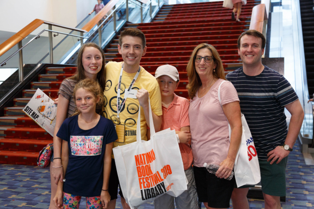 Eine Gruppe von Menschen, die vor einer Rolltreppe stehen und Taschen halten, vor einer Treppe, Geländern und einer Wand im Hintergrund.