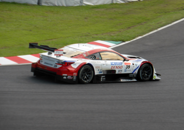 Ein Sportwagen mit Text und Zahlen, der bei den 24 Stunden von Le Mans teilnimmt, fährt auf einer Rennstrecke mit Gras änderseits und einem Zaun im Hintergrund.