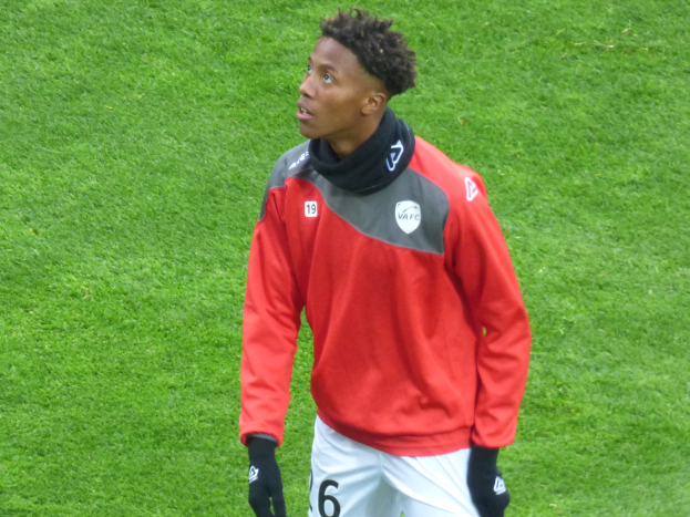 Ein junger Mann in einer roten Jacke und weißen Shorts steht auf einem Fußballfeld und schaut rechts mit entschlossener Miene, wobei er Baseball-Handschuhe trägt.