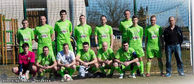 Eine Gruppe von Männern in grünen T-Shirts steht und hockt auf einem Fußballfeld mit einem Ball davor, ein Torpfosten rechts und ein Hintergrund aus Bäumen, Gebäuden, Pfählen, Fahrzeugen und Himmel, mit Text, der Zahlen unten links auflistet.