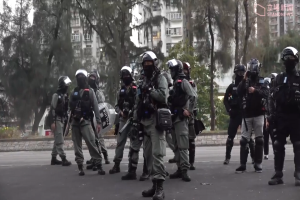 Eine Gruppe von Polizisten in Einsatzausrütstüßtz, darunter Helme und mit Waffen, geht eine Straße mit Bäumen, Gebäuden und einem klaren Himmel im Hintergrund entlang; ein Wasserzeichen ist auf der rechten Seite sichtbar.