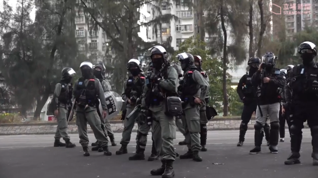 Eine Gruppe von Polizisten in Einsatzausrütstüßtz, darunter Helme und mit Waffen, geht eine Straße mit Bäumen, Gebäuden und einem klaren Himmel im Hintergrund entlang; ein Wasserzeichen ist auf der rechten Seite sichtbar.