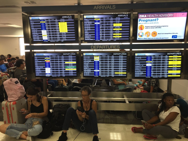 Eine Gruppe von Menschen sitzt auf dem Boden eines Flughafens, einige halten Handys, mit verstreuten Taschen und Bildschirmen, die Zika-Virus-Informationen anzeigen, im Hintergrund.