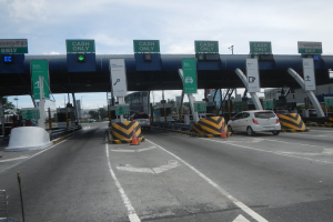 Mautstation mit darunter fahrenden Autos, mit Verkehrskegeln, Absperrpollern, Hinweistafeln, einem Bogen, Laternenmasten, Bäumen, Gebäuden und einem bewölkten Himmel im Hintergrund.