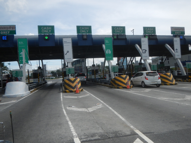 Mautstation mit darunter fahrenden Autos, mit Verkehrskegeln, Absperrpollern, Hinweistafeln, einem Bogen, Laternenmasten, Bäumen, Gebäuden und einem bewölkten Himmel im Hintergrund.