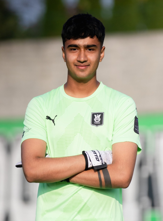 Ein junger Mann in grünem T-Shirt steht auf einem Fuballfeld und verschräkelt die Arme, lächelt, mit einem unscharfen Hintergrund.