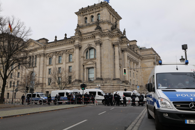 Eine Gruppe von Polizeibeamten steht vor dem Reichstaggebäude in Berlin, Deutschland, mit Fahrzeugen, einem Zaun, Verkehrsampeln, Laternen, Bäumen und Flaggen im Hintergrund, bei klarem Himmel.