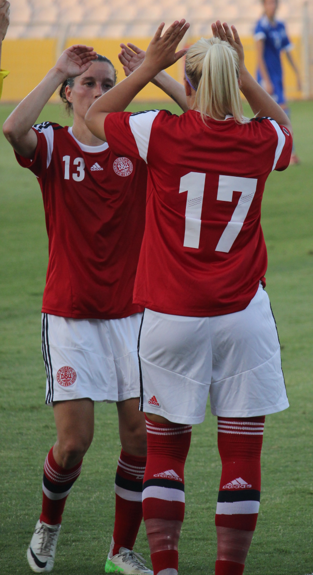 Zwei Frauen in roten T-Shirts und weißen Shorts stehen auf einem Fuballfeld und klatschen jubelnd in die Hände, mit einer Person in einem blauen Kleid, einer gelben Tafel und einigen Stühlen im Hintergrund.