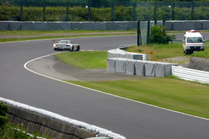 Ein Rennwagen fährt eine Rennstrecke entlang, im Hintergrund ist ein Polizeiwagen zu sehen, umgeben von Grünflächen und Absperrungen, mit Text unten.
