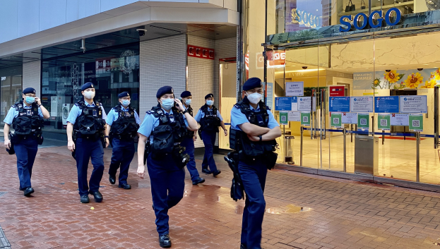 Eine Gruppe von Polizisten in Gesichtsmasken, die vor einem Gebäude mit Glaswänden und -türen eine Straße entlanggehen, mit einem Geschäft auf der rechten Seite, das Plakate an seinen Glaswänden zeigt.