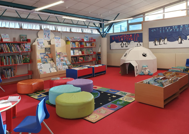 Eine Kinderbibliothek voller Bücher, Möbel, Lehrmittel und natürlichem Licht durch große Fenster.