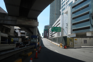 Stadtstraße mit Verkehrskegeln, Fahrzeugen, einer Brücke links, Gebäuden mit Namensschildern, Laternen und einem Himmel im Hintergrund.