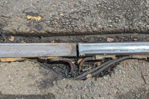 Nahaufnahme eines Metallkanals auf der Straßenseite mit daran befestigten Drähten und verstreuten trockenen Blättern.