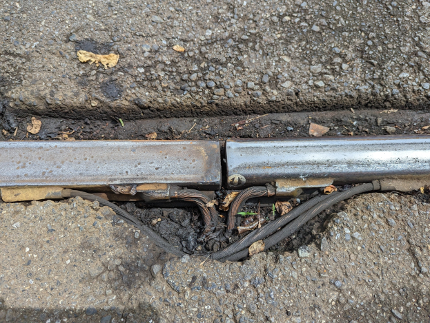 Nahaufnahme eines Metallkanals auf der Straßenseite mit daran befestigten Drähten und verstreuten trockenen Blättern.