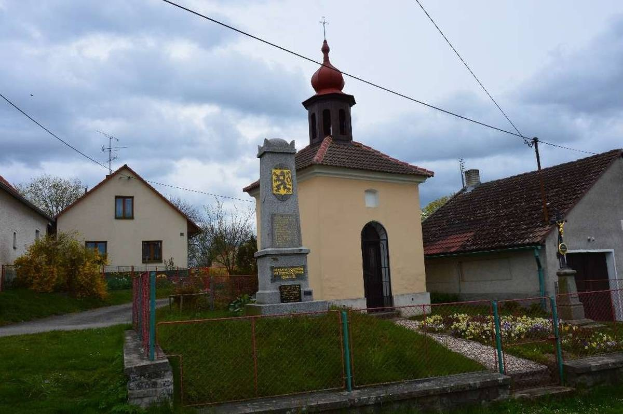 Kleine Kirche mit einem Glockenturm, identifiziert als die Kirche des Heiligen Grabes, umgeben von Häusern, einem Zaun, Gras, blühenden Pflanzen, einem Pfad, Bäumen, Strommasten mit Drähten und einem bewölkten Himmel.
