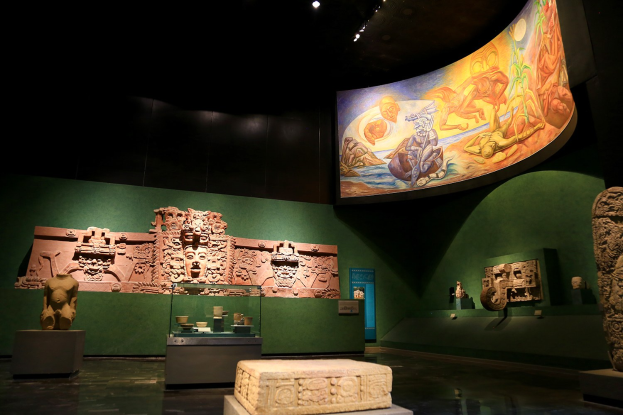 Ausstellungen mit Skulpturen, Artefakten auf Tischen, einer Texttafel, einer Wandmalerei und Deckenleuchten im Mexiko-Stadt-Museum der Anthropologie.