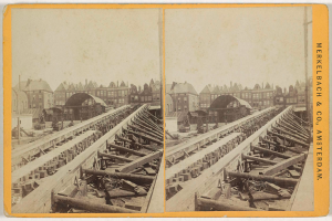 Ein altes Schwarz-Weiß-Foto einer Bahntrasse mit Gebäuden im Hintergrund, Text auf der rechten Seite mit 'Merkelbach & Co, Co. Amsterdam' und verstreuten Holzobjekten im Vordergrund.