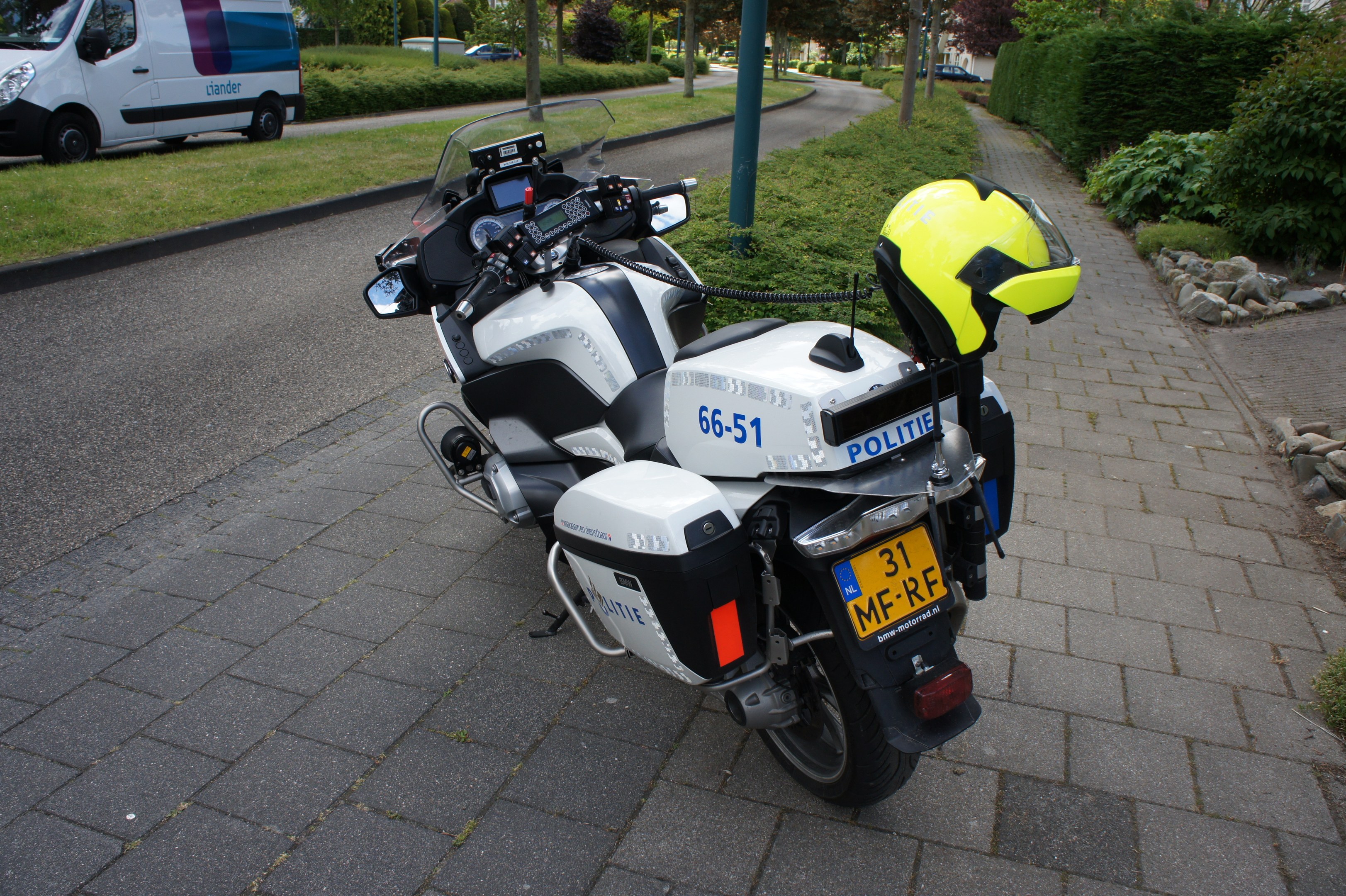 Ein Polizeimotorrad steht am Straßenrand mit einem Helm darauf, umgeben von Bäumen, Pfählen, Gebäuden, Pflanzen, Gras, Steinen und anderen Fahrzeugen.