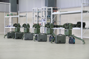 Eine Gruppe von Militärrobotern ist auf einem Boden positioniert und von Koffern und anderen Gegenständen auf Gestellen umgeben, mit einer Wand und einem Geländer im Hintergrund.