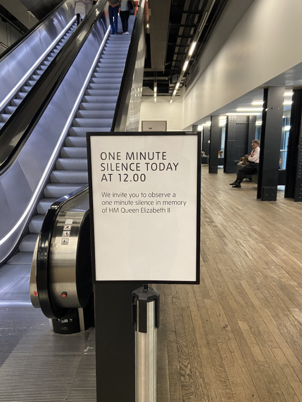 Eine Rolltreppe im Flughafen mit einem Schild, auf dem "Eine Minute Schweigen heute" steht, ein paar Menschen darauf und Leuchten an der Decke im Hintergrund.