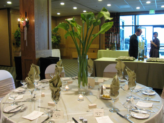 Hotel-Speisesaal mit einem weißgedeckten Tisch, auf dem Teller, Besteck und ein Glas stehen, umgeben von Stühlen, einem grünen Tisch im Hintergrund, einem Mann in einem schwarzen Blazer, einem stehenden Mädchen, einer Tür mit der Aufschrift "Ausgang" und zwei Männern dahinter.