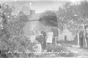 Ein altes Schwarz-Weiß-Foto einer Familie, die vor einem Haus steht, mit einer Frau, die ein Baby hält, umgeben von anderen Familienmitgliedern, Bäumen und einer Hütte im Hintergrund, und Text am unteren Rand.