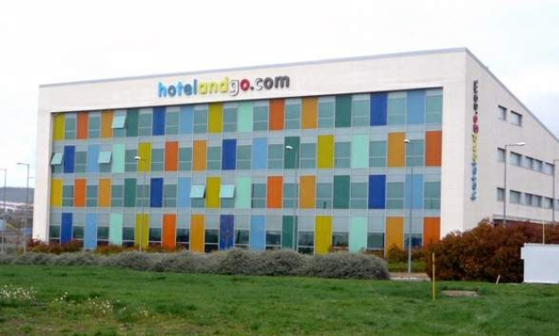 Großes modernes Hotelgebäude mit zahlreichen Fenstern, umgeben von Grünflächen, einem Zaun, Straßenlaternen, geparkten Fahrzeugen, fernen Hügeln und einem bewölkten Himmel; das Gebäude trägt die Aufschrift "hotel und go".