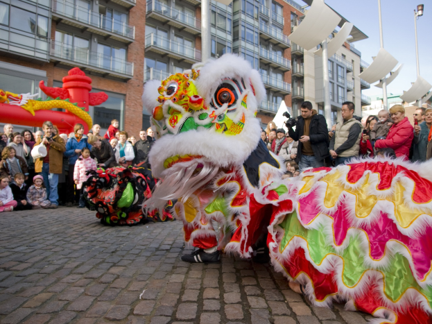 Ein lebendiges chinesisches Neujahrsfest in Amsterdam mit einem Löwen tanzen im Vordergrund und einer Menge Schaulustiger, einige halten Kameras, vor einem Hintergrund aus Gebäuden, Laternenmasten und einem klaren blauen Himmel.