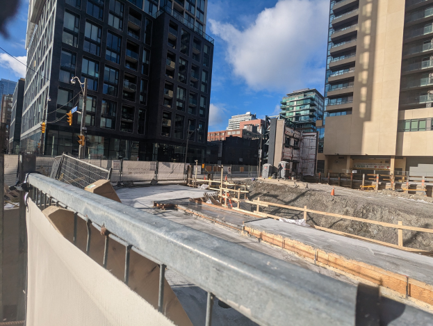 Baustelle in Toronto, Ontario mit einem großen Bauwerk im Hintergrund, umgeben von Absperrungen, Pfählen, Verkehrszeichen und Schildern unter einem bewölkten Himmel.