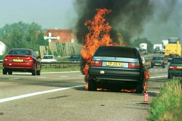 Ein Auto in Flammen an der Straße, umgeben von anderen Fahrzeugen, mit Bäumen, Gebäuden und einem klaren blauen Himmel im Hintergrund; ein Feuerlöscher ist auf der rechten Seite zu sehen.