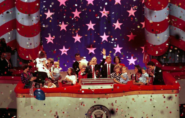 Eine Gruppe von Menschen auf einer Bühne mit einem Rednerpult, das ein Logo, Luftballons und Konfetti zeigt, mit Sternen und einem Vorhang im Hintergrund.