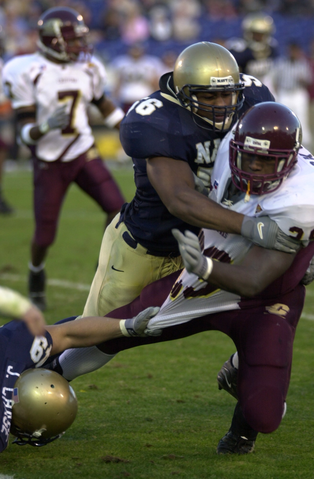 Zwei Fussballspieler in Helmen und Uniformen kämpfen auf dem Feld, während andere zusehen.