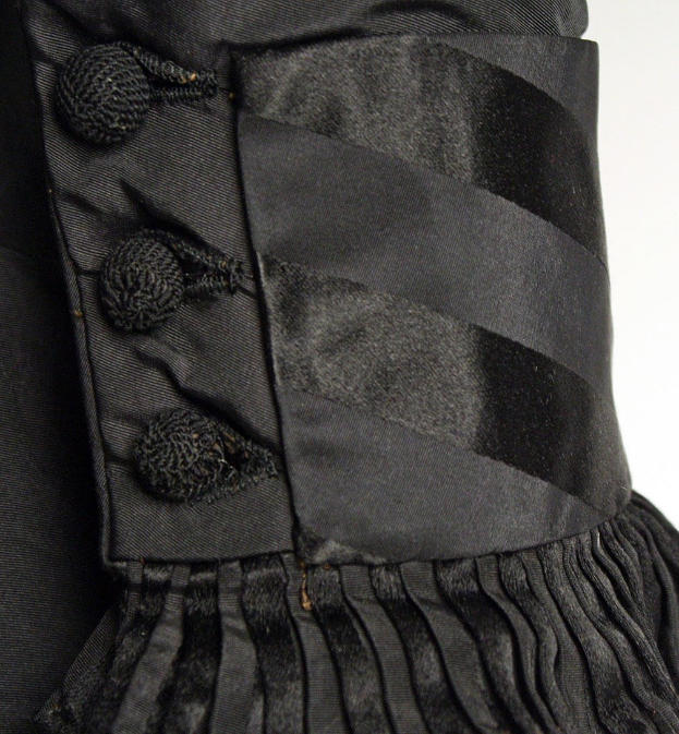 Nahaufnahme eines schwarzen, knielangen Kleides mit silbernen Knöpfen in symmetrischem Muster, mit einer eng anliegenden Taille, hohem Bund und plissierter Röcke vor einem weißen Hintergrund.