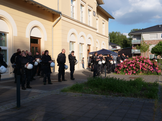 Eine Gruppe von Polizisten steht vor einem Gebäude mit Fenstern und Türen, hält Helme in den Händen, mit einem Zelt in der Nähe, umgeben von Pflanzen mit Blumen, Gras, Bäumen und einem Straßenpfahl unter einem bewölkten Himmel.