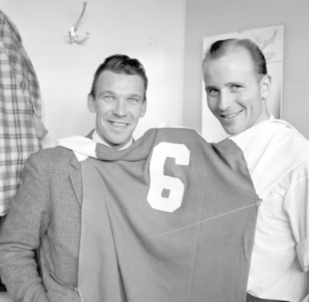 Zwei Männer, die strahlend ein Fußballtrikot hochhalten, mit einer Wandtafel und einem Haken im Hintergrund, in Schwarz-Weiß.
