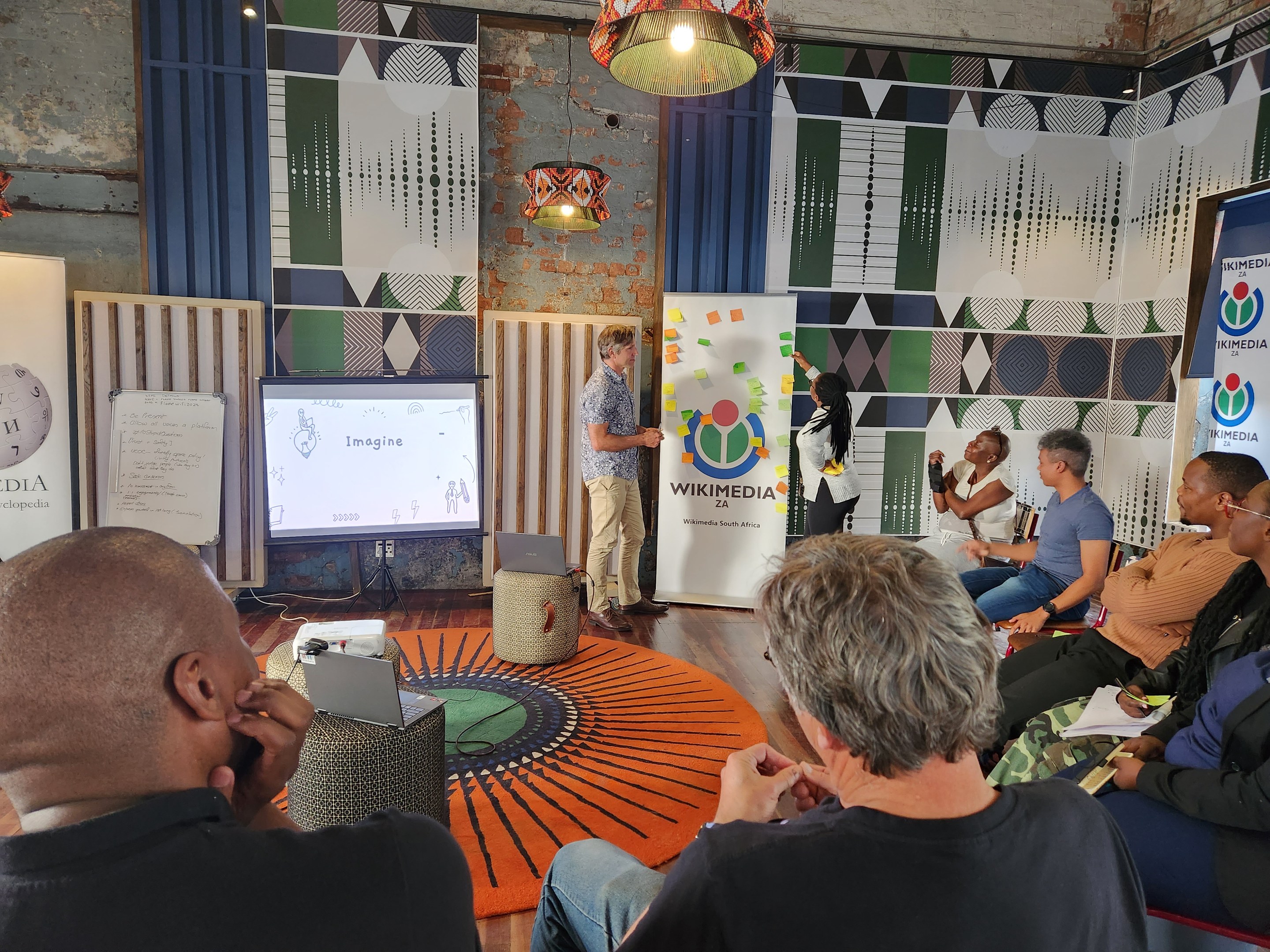 Eine Gruppe von Menschen, die in Stühlen vor einem Projektionsschirm sitzen, mit einem Mann, der vorne steht und ein Mikrofon hält; auf der linken Seite ein Tisch mit einem Laptop und anderen Gegenständen, und im Hintergrund Schilder mit dem Text "Wikimedia 2a - die Zukunft des digitalen Marketings", eine Blumenvase und zwei Hängelampen.