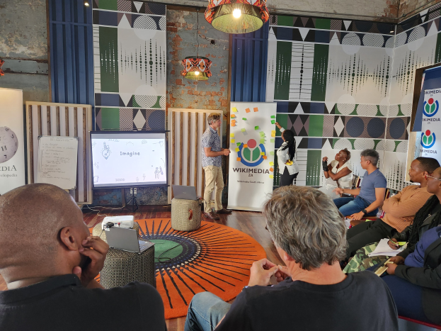 Eine Gruppe von Menschen, die in Stühlen vor einem Projektionsschirm sitzen, mit einem Mann, der vorne steht und ein Mikrofon hält; auf der linken Seite ein Tisch mit einem Laptop und anderen Gegenständen, und im Hintergrund Schilder mit dem Text "Wikimedia 2a - die Zukunft des digitalen Marketings", eine Blumenvase und zwei Hängelampen.