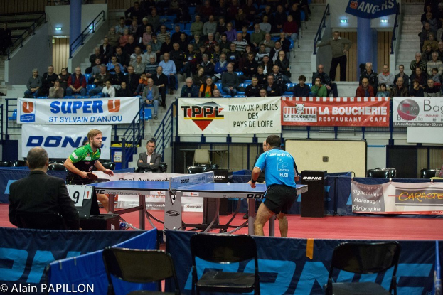 Indoor-Tischtennis-Match mit zwei Spielern und einigen stehenden Zuschauern, sitzendem Publikum, leeren Stühlen und Werbeplakaten in einer Stadionatmosphäre.