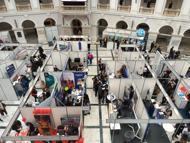 Eine große Gruppe von Menschen versammelte sich in einem Raum, der mit Ständen, Bannern, Tischen, Stühlen und Luftballons gefüllt war, mit einem Gebäude im Hintergrund, das Bögen, Säulen, Geländer und einen Bildschirm aufweist.